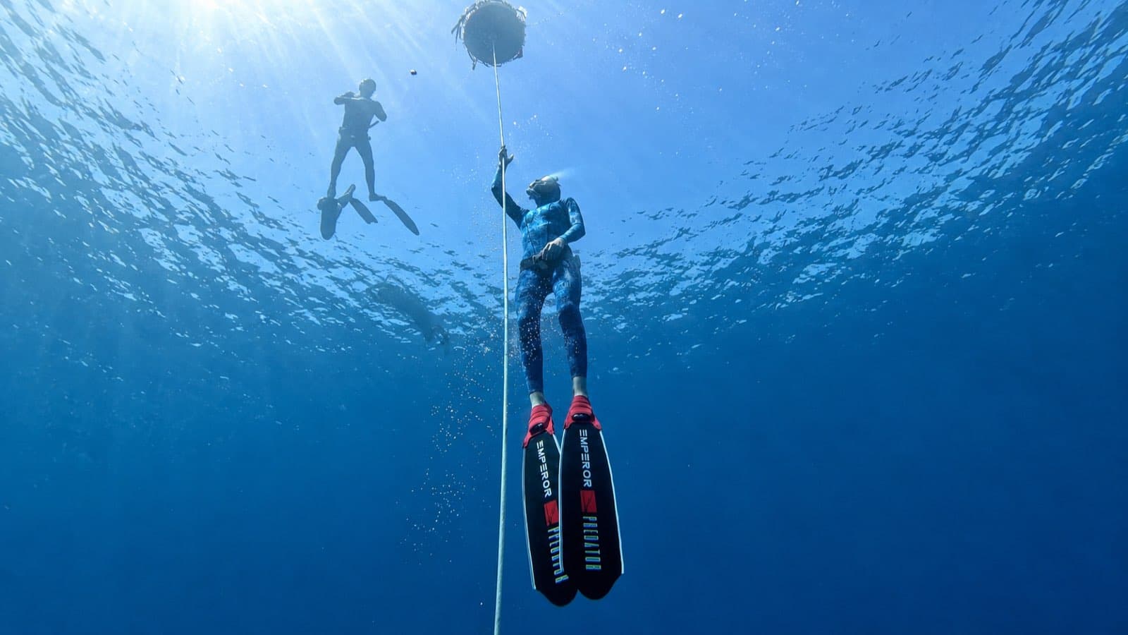A freediver in Predator fins holding a dive line below the surface, sun rays through the water